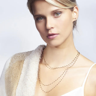 A woman wearing a white dress and a beige coat with a gold necklace, Gigi Clozeau Classic Necklace Quartz & Yellow Gold, layered over the white neckline.