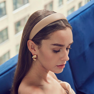 Woman wearing a beige headband Lele Sadoughi Khaki Faux Leather Bessette Headband and gold earrings with a blurred background